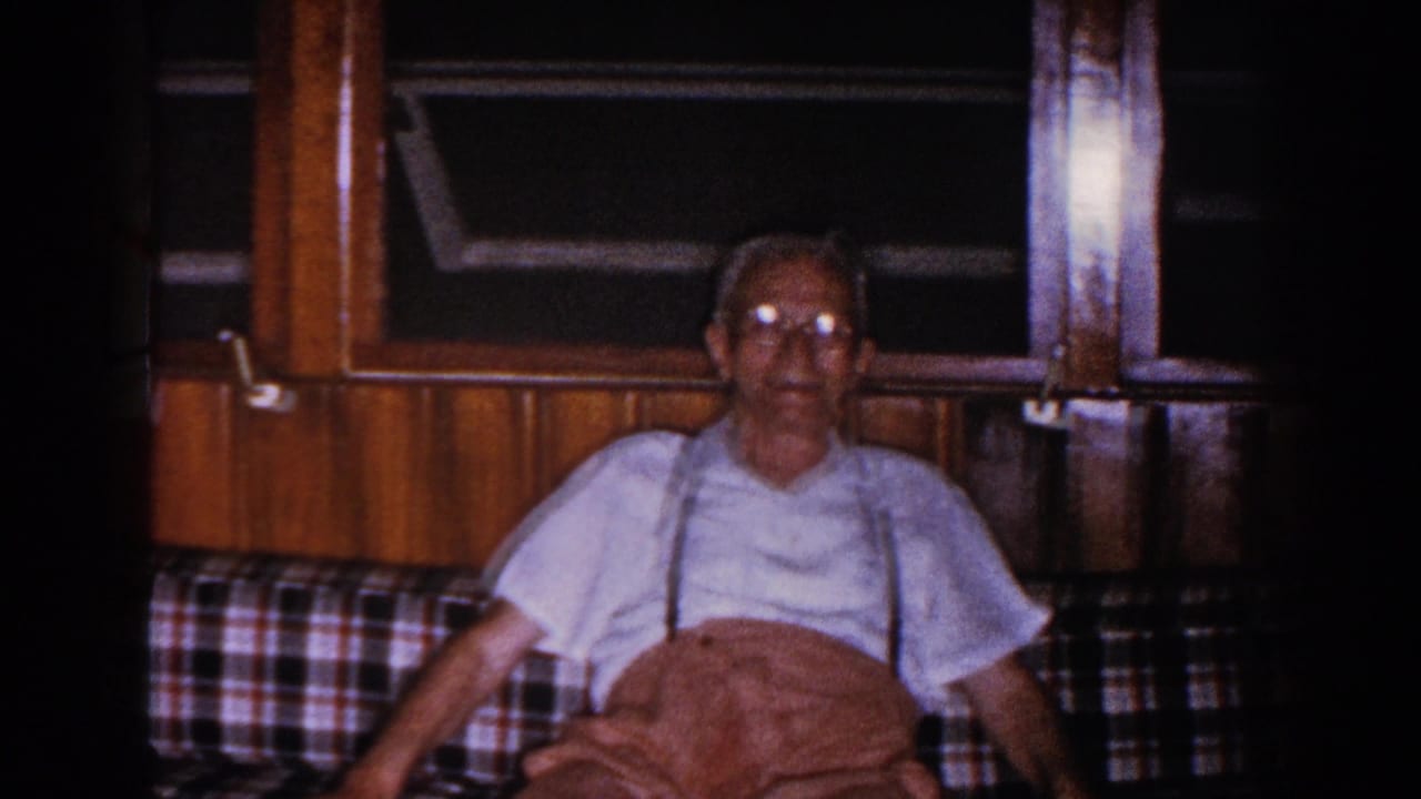 Older man in suspenders sits on a plaid couch in a wood-paneled room.