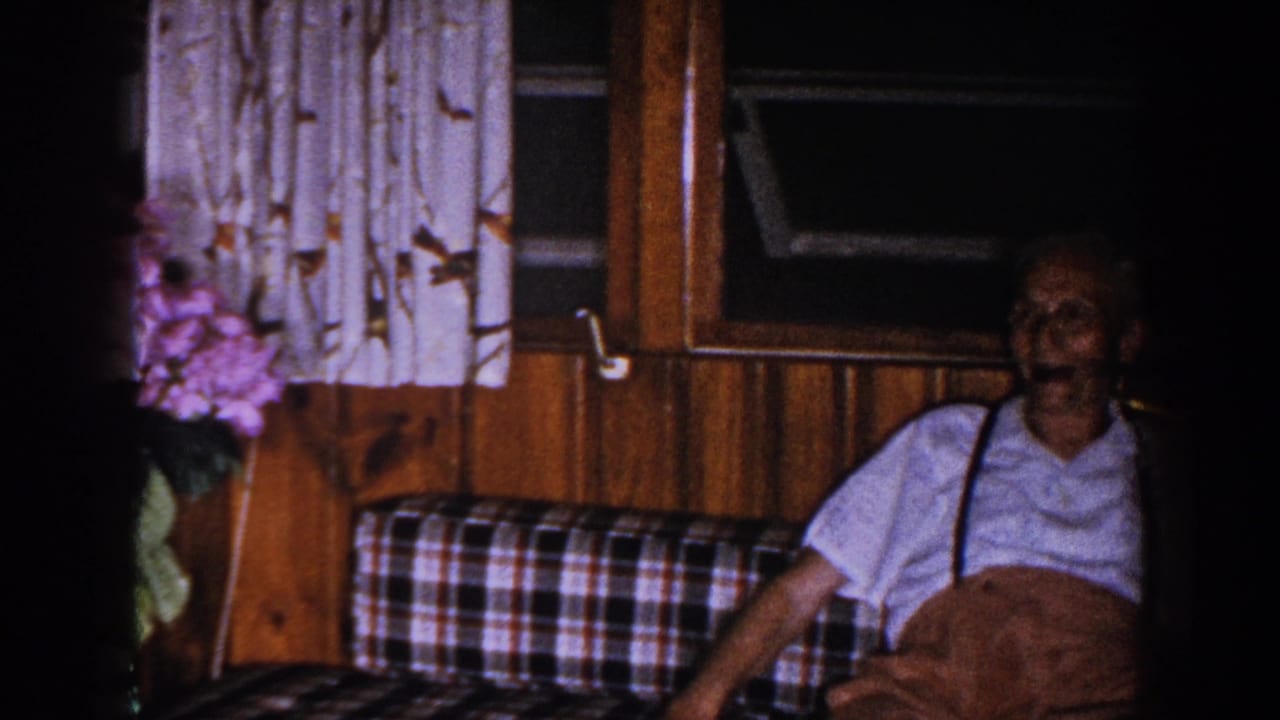 Older man sits partly in shadow beside a window and patterned curtains.