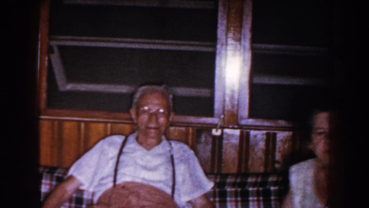 Older man seated on a couch with a woman partly visible at the edge of the room.
