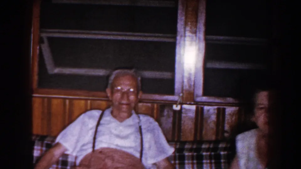 Older man remains seated on the plaid couch while a woman appears partly at frame edge in the 1960 Brighton home interior.