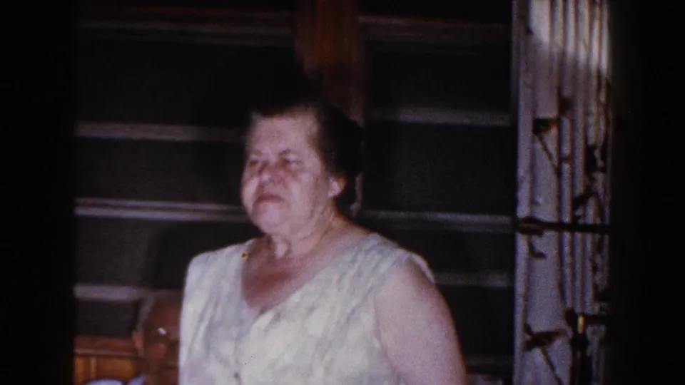 Older woman stands near the couch inside the Brighton, Michigan home, with a seated man partly visible behind her.