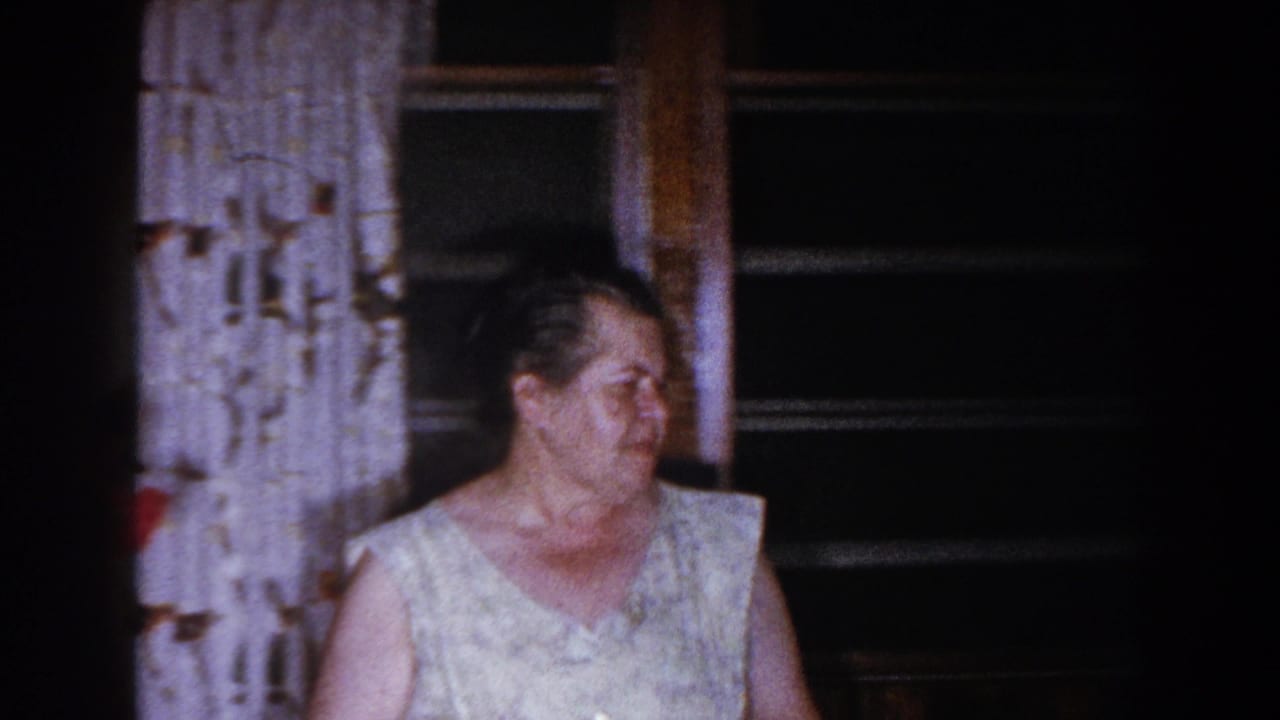 Older woman in a light sleeveless dress standing near patterned curtains and a dark window.