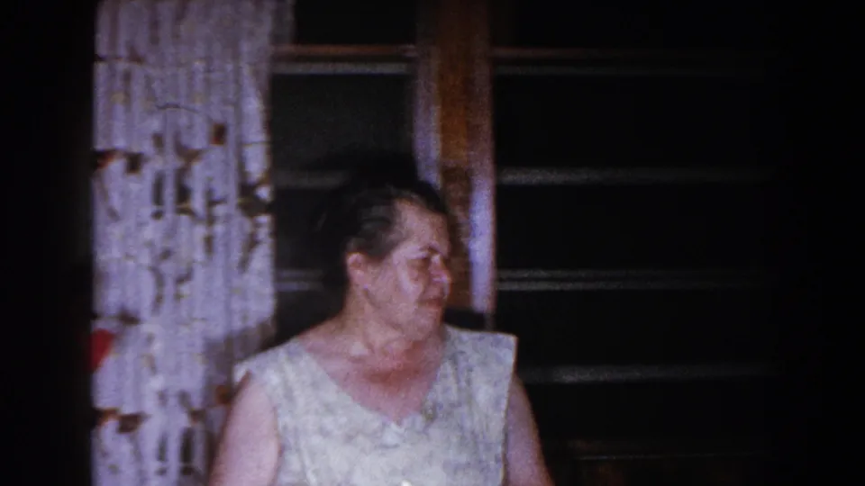 Older woman in a light sleeveless dress inside a Brighton, Michigan living room, near patterned curtains and a dark window.