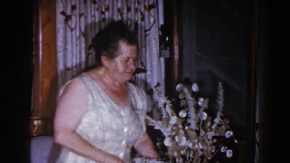 Older woman in a light dress looks down beside a pale floral or dried-stem arrangement in the Brighton living room.