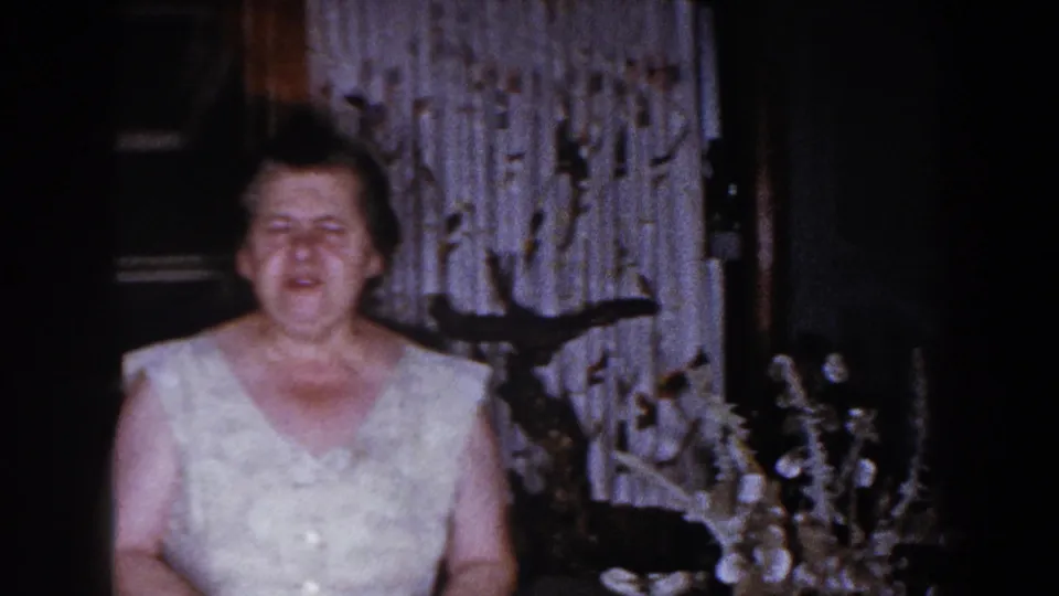 Older woman in a sleeveless white dress appears to face the camera inside a dim Brighton, Michigan home interior in 1960.