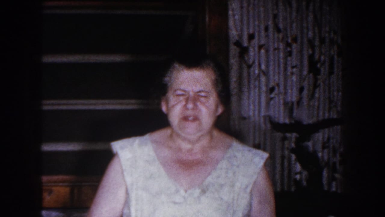 Older woman in a light sleeveless dress centered in a dark room with curtains and shelves behind her.