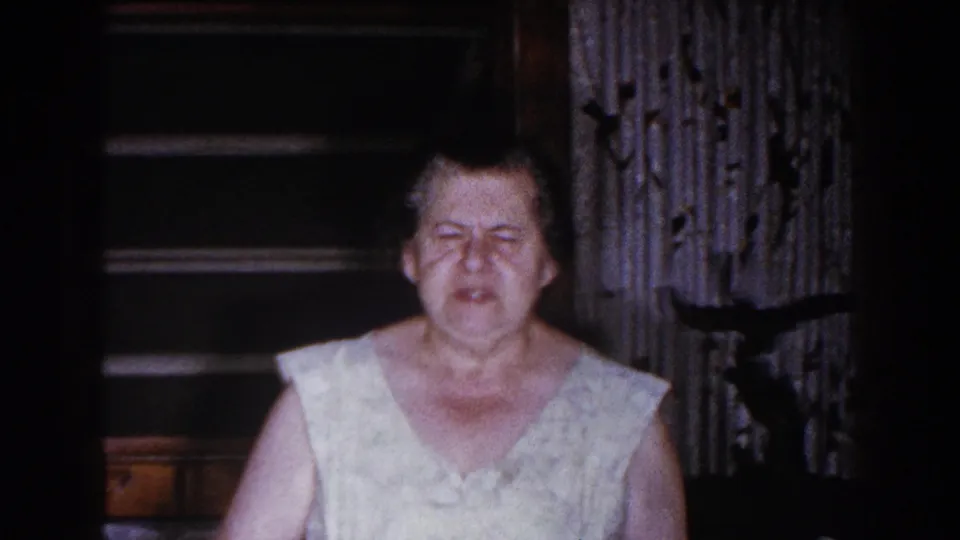 Older woman in a light sleeveless dress stands or moves toward the camera in a dark 1960 home interior in Brighton, Michigan.