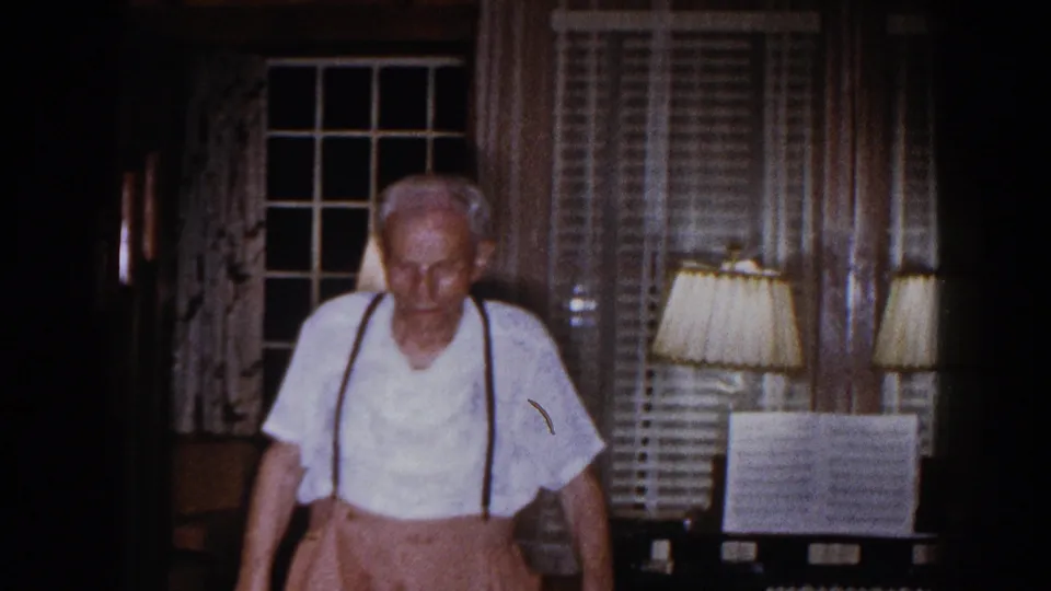 Older man in a white shirt with suspenders walks toward the camera inside a 1960 Brighton, Michigan home interior.
