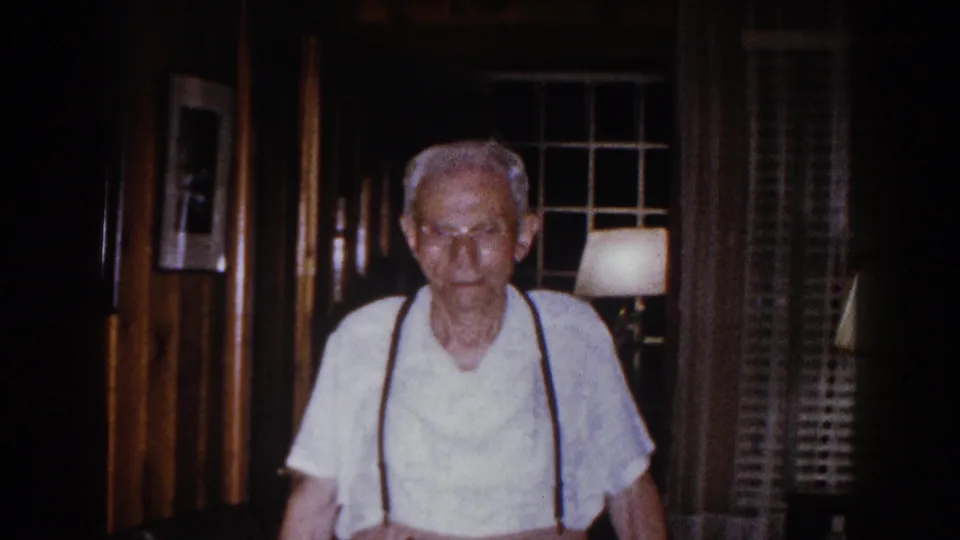 Older man wearing a white shirt and dark suspenders approaches the camera in a wood-trimmed 1960 home interior.