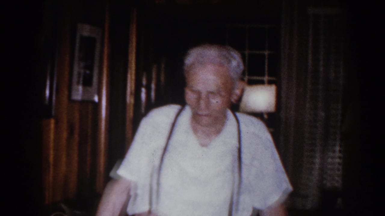 Elderly man in a light shirt and dark suspenders walks through a dim Brighton, Michigan living room in 1960 home-movie footage.
