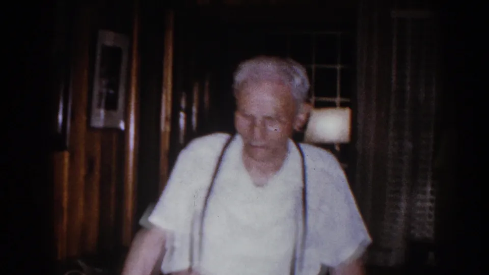 Elderly man in a light shirt and dark suspenders walks through a dim Brighton, Michigan living room in 1960 home-movie footage.