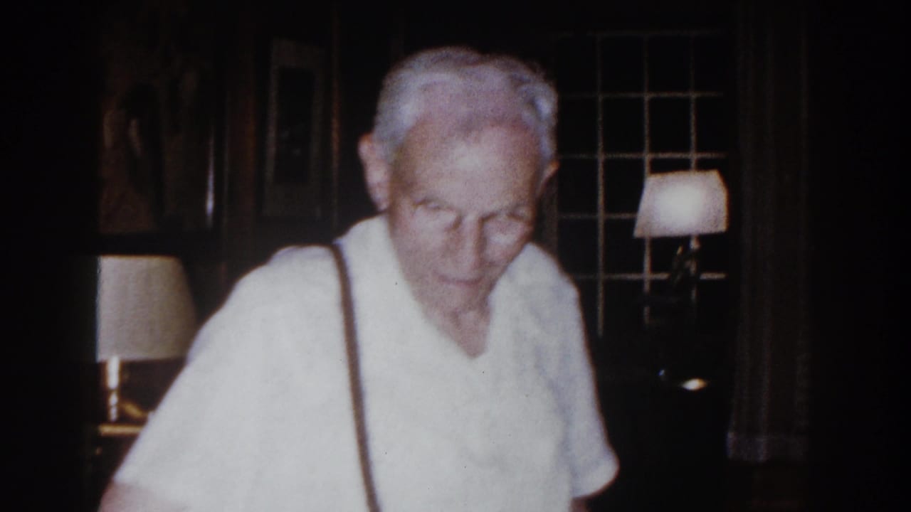 Elderly man faces the camera near lit lamps and dark windows.