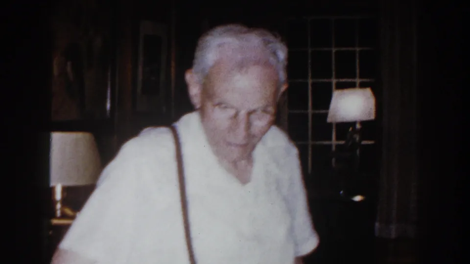 Elderly man with white hair and suspenders faces the camera inside the Brighton, Michigan living room, with lamps and windows behind him.
