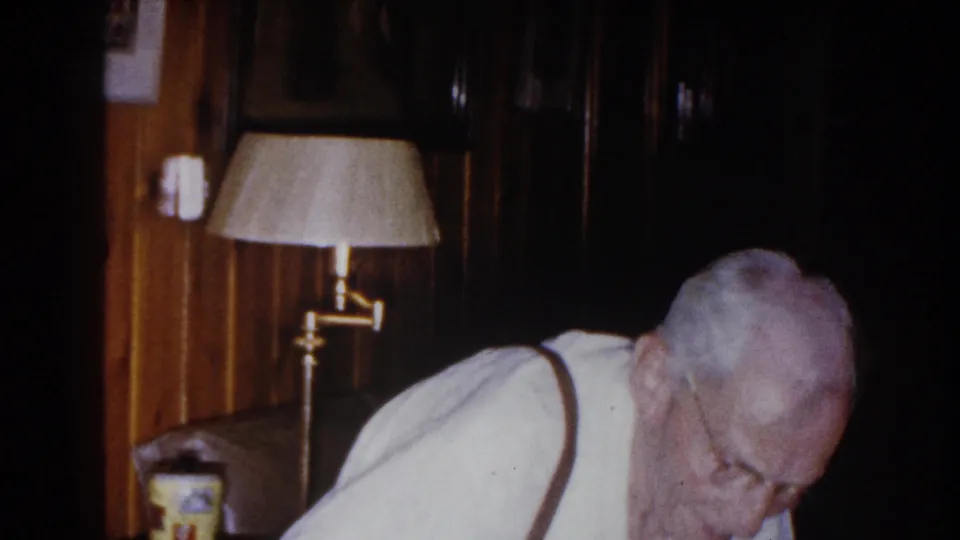 Elderly man wearing glasses bends forward near a shaded floor lamp and wood-paneled wall in 1960 Brighton, Michigan home footage.