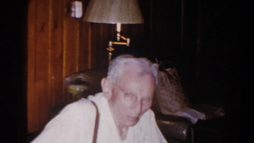 Elderly man in suspenders leans forward in a warm, grainy Brighton, Michigan living room frame with a chair and lamp visible.