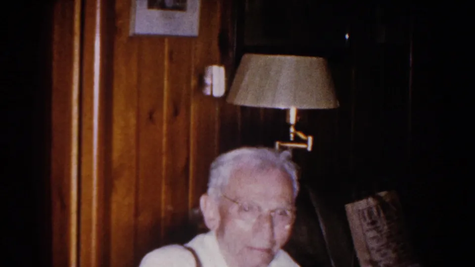In a 1960 Brighton, Michigan home interior, an elderly man with glasses sits below a wall lamp amid wood paneling and framed pictures.