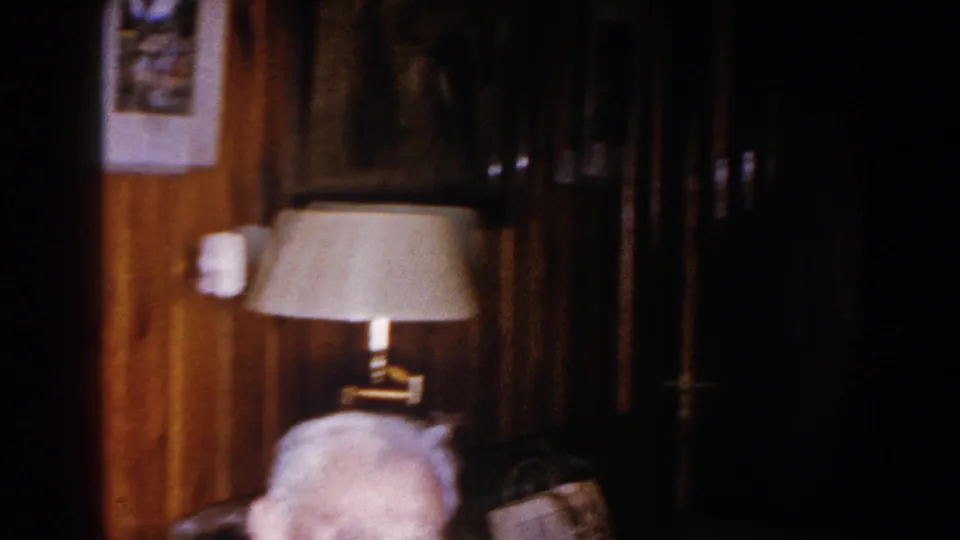 The camera catches the top of an elderly man's head below a shaded lamp, with dark wood paneling and framed decor visible.