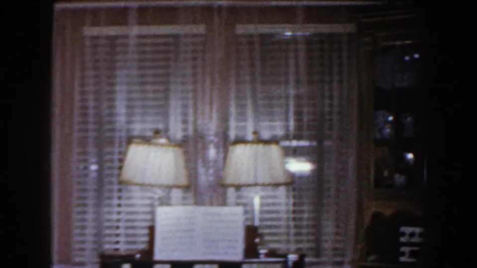 A piano with open sheet music stands before curtained windows and blinds, with lamps reflected or placed nearby in the dim room.