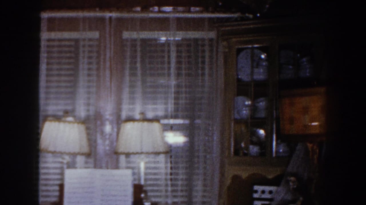 1960 Brighton, Michigan home-movie frame showing a dim family room with double windows, lamps, piano, and china cabinet.
