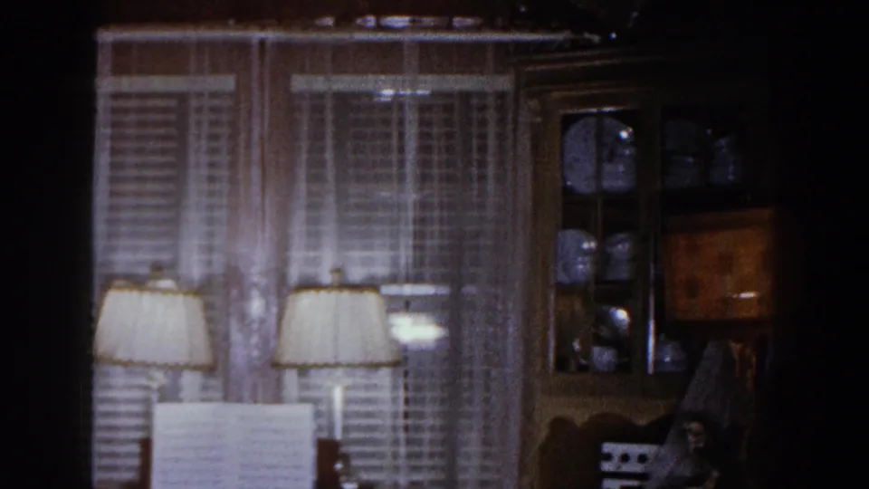 1960 Brighton, Michigan home-movie frame showing a dim family room with double windows, lamps, piano, and china cabinet.