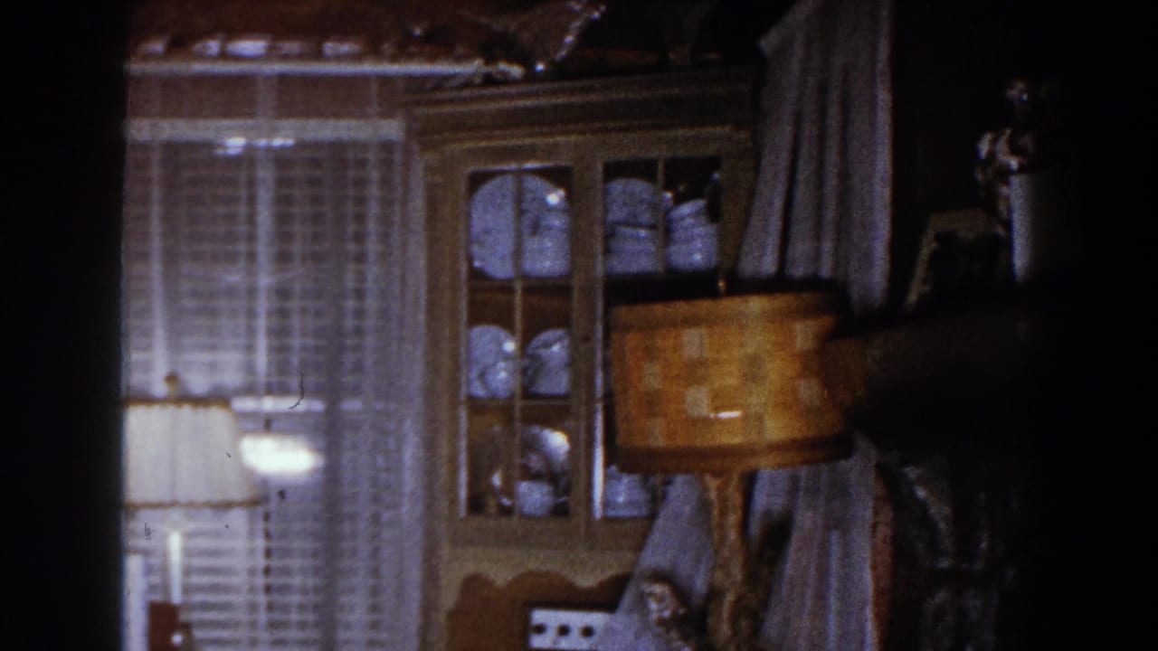 1960 Brighton, Michigan interior frame with a lit lamp, curtained windows, china cabinet, and dark edge from the room scan.