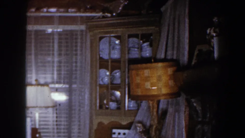 1960 Brighton, Michigan interior frame with a lit lamp, curtained windows, china cabinet, and dark edge from the room scan.