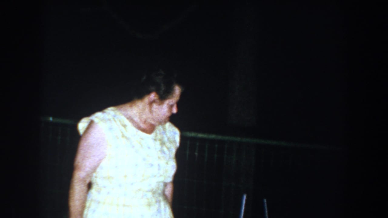 In Brighton, Michigan 1960 home-movie footage, an older woman in a light patterned dress looks down near a fence at night.
