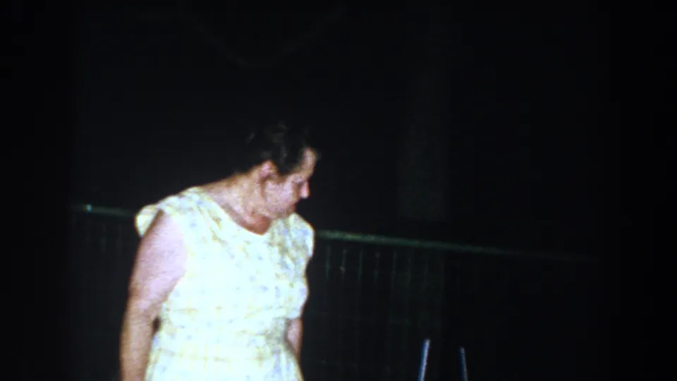 In Brighton, Michigan 1960 home-movie footage, an older woman in a light patterned dress looks down near a fence at night.