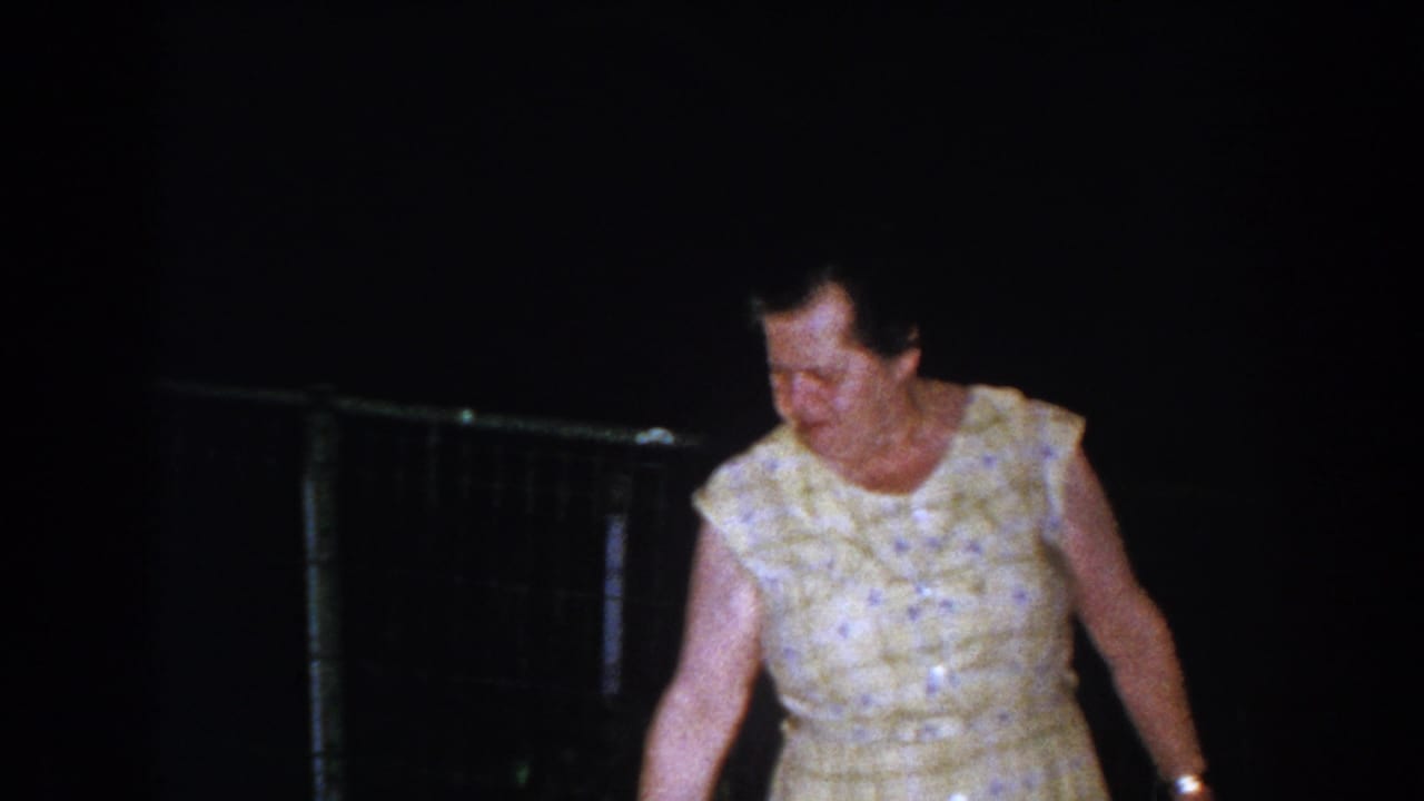 Older woman in a yellow patterned dress walking in a dark backyard.