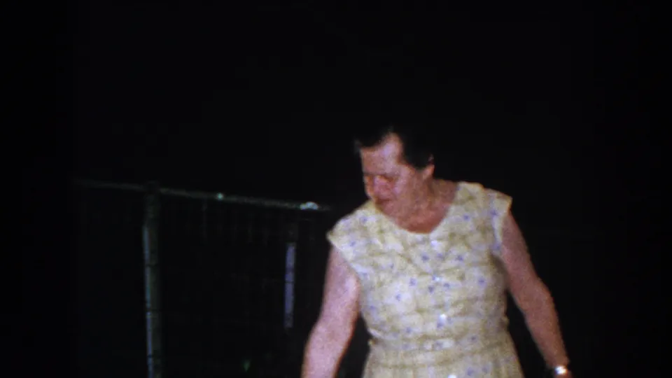 Older woman in a yellow patterned dress walks through a dim backyard in Brighton, Michigan, circa 1960.