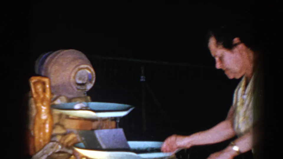 Older woman in a yellow patterned dress bends beside the backyard fountain and reaches into the water.