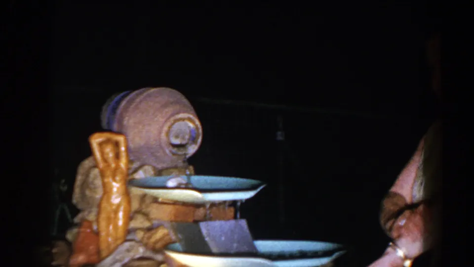 1960 Brighton, Michigan 8mm frame showing a ceramic and stone-style fountain beside a partially visible adult in a dark outdoor setting.