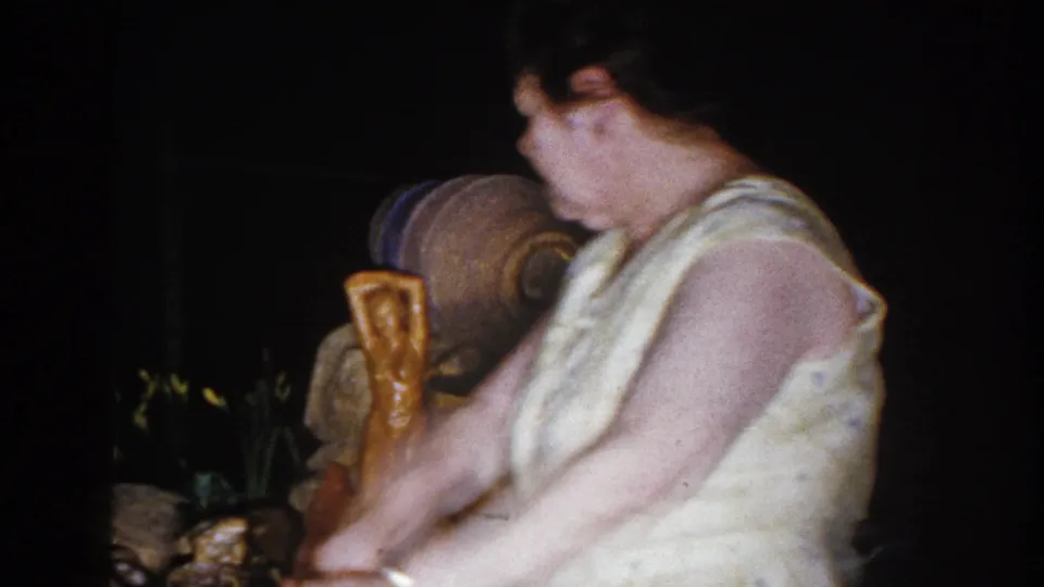 1960 Brighton, Michigan home-movie frame of a woman in a light dress looking toward a small artificial yard fountain.
