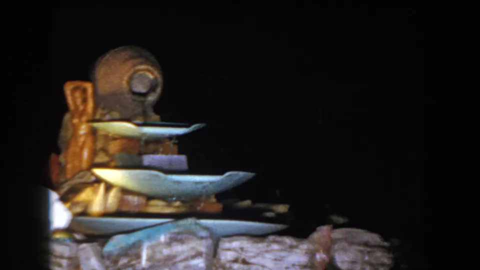 1960 Brighton, Michigan home-movie frame showing a small tiered fountain with blue basins, brown wood, and a stone base at night.