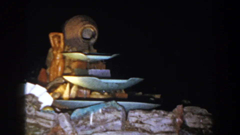 The 8mm frame holds on the outdoor fountain, showing stacked blue-tinted basins, carved brown wood, and rocks below.