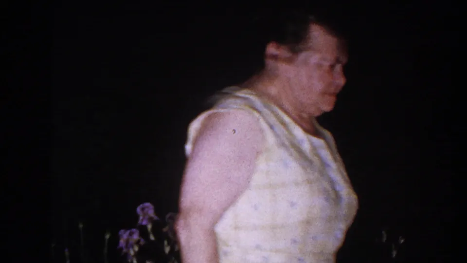 An older woman in a light sleeveless top walks through a dark Brighton, Michigan garden past purple irises in 1960 8mm footage.