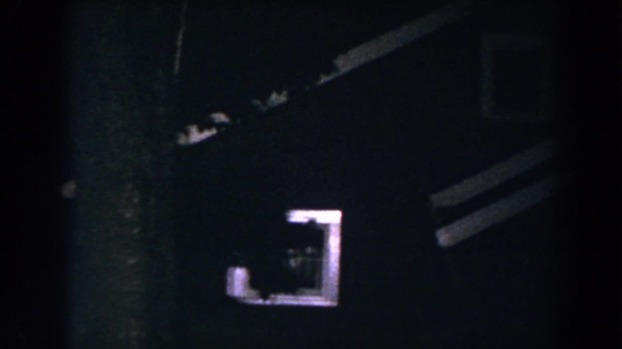 1960 8mm home-movie frame from Brighton, Michigan showing a dark house exterior at night with one glowing window and partial rooflines.