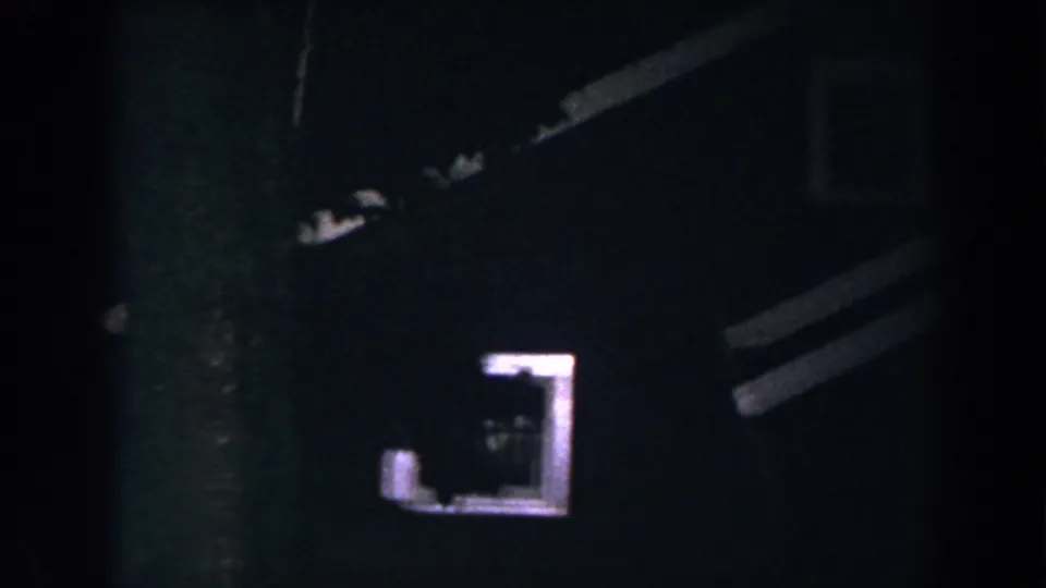1960 8mm home-movie frame from Brighton, Michigan showing a dark house exterior at night with one glowing window and partial rooflines.