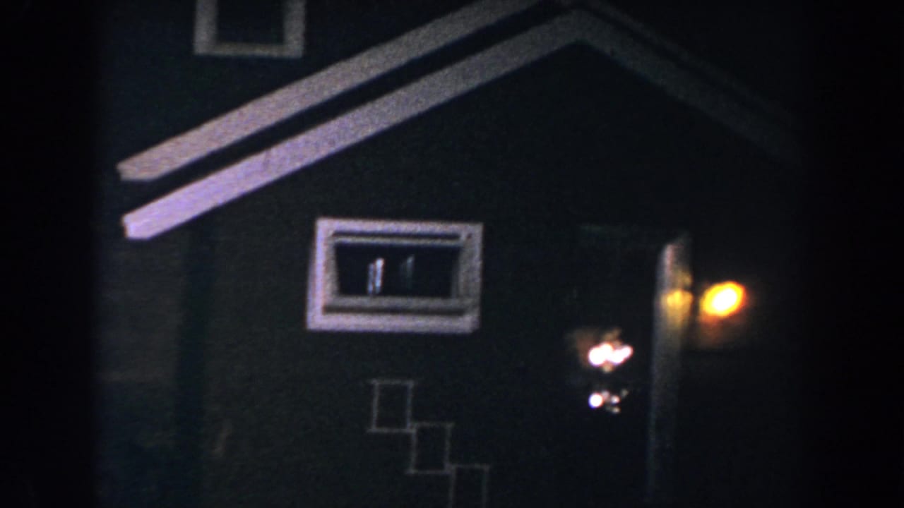 1960 8mm frame in Brighton, Michigan showing a house facade at night with small windows, roof trim, and a warm porch light by the entrance.