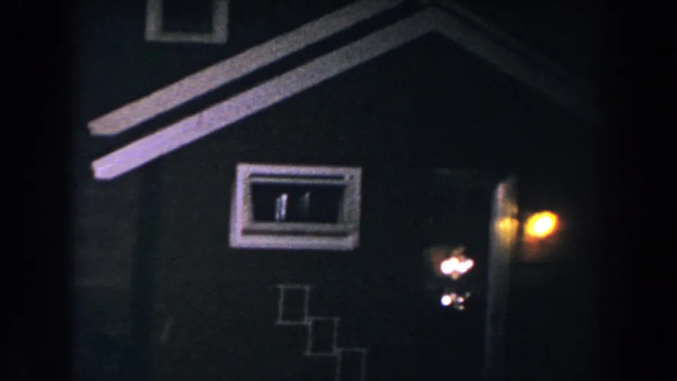 1960 8mm frame in Brighton, Michigan showing a house facade at night with small windows, roof trim, and a warm porch light by the entrance.