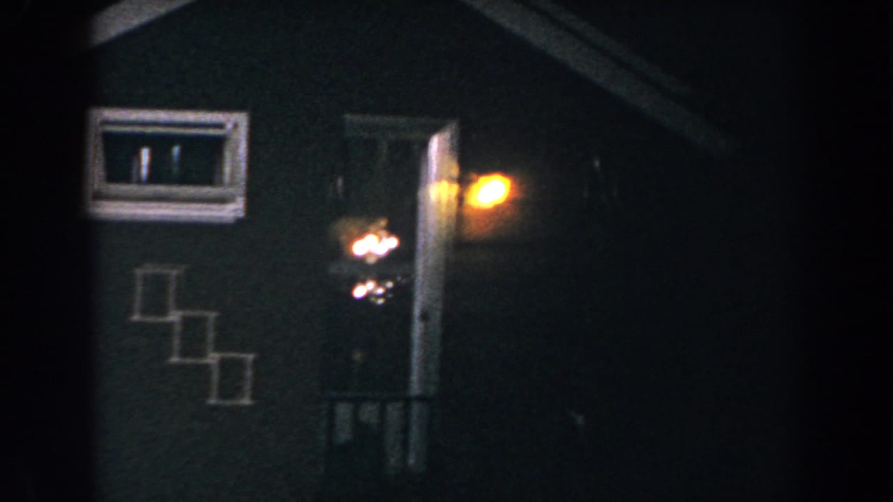 1960 8mm home-movie frame from Brighton, Michigan showing a dark residential entrance with a glowing porch light, lit window, and exterior steps.