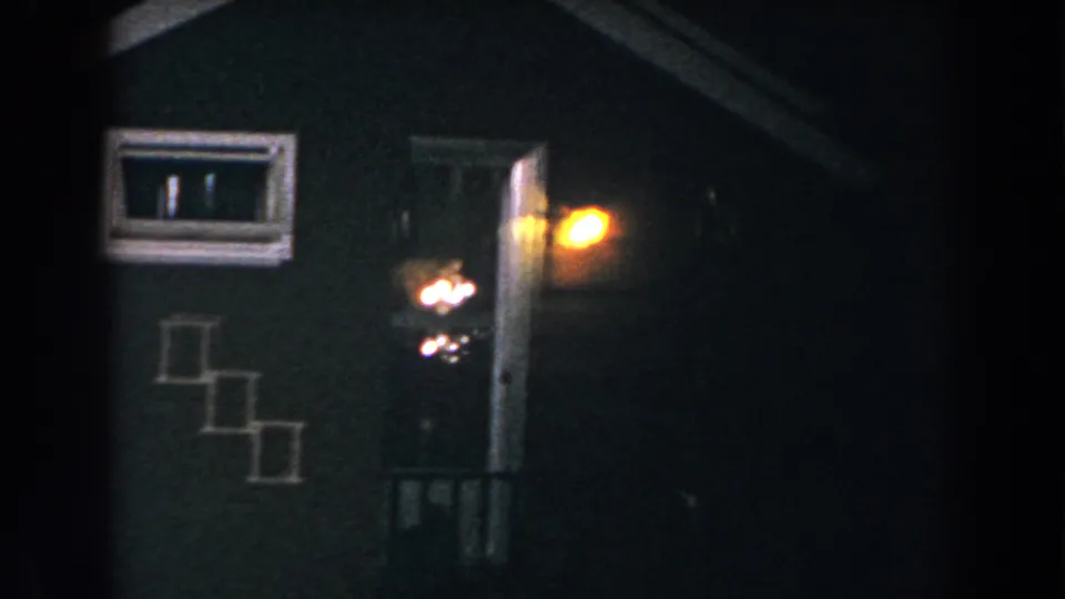 1960 8mm home-movie frame from Brighton, Michigan showing a dark residential entrance with a glowing porch light, lit window, and exterior steps.