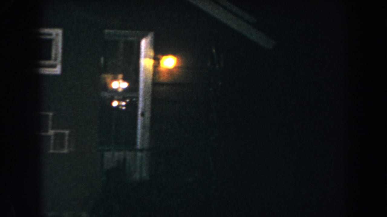 1960 8mm frame in Brighton, Michigan showing the side of a dark house at night, with a warm porch light and doorway area partly obscured by darkness.