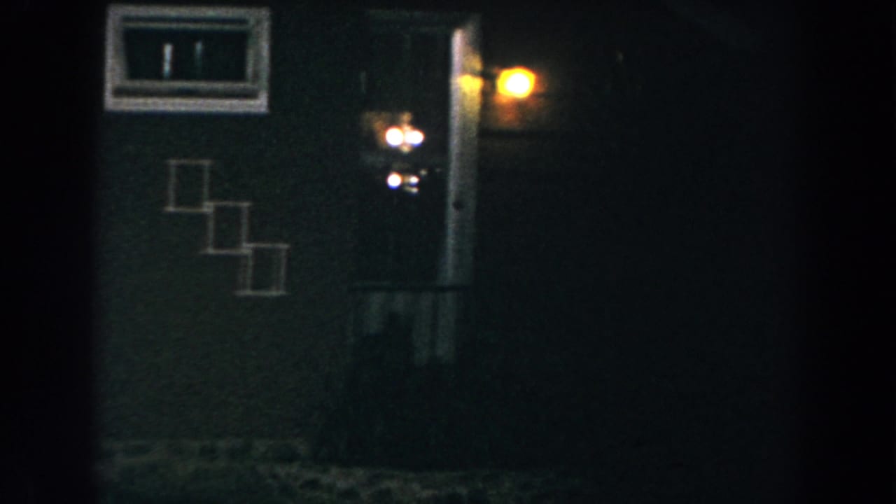 Dark house exterior with a small window, doorway light, and part of a wooden fence.