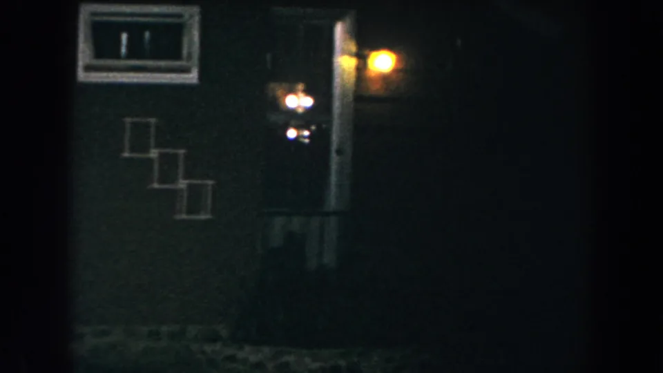 1960 Brighton, Michigan 8mm frame showing a dim rural house exterior with a small window, lit doorway area, and fence in the foreground.