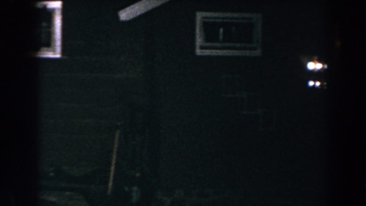 Dark wall of a house with a small window, exterior light, and shadowed yard objects.