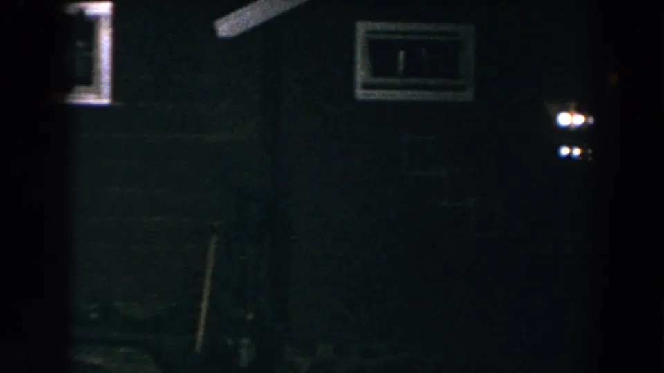 1960 Brighton, Michigan 8mm frame of a dark rural exterior with a small window, bright exterior light, and indistinct yard objects.
