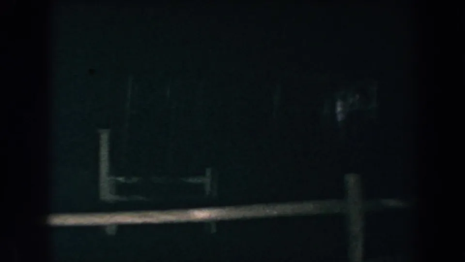 Low-light 8mm frame from Brighton, Michigan, with fence rails, posts, and a faint distant flare in darkness.