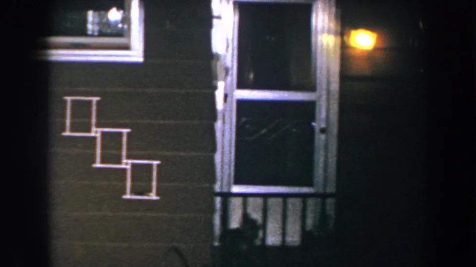 1960 Brighton, Michigan home-movie frame showing a brown residential wall at night with white-trimmed steps, a screen door, porch railing, and a yellow exterior light.