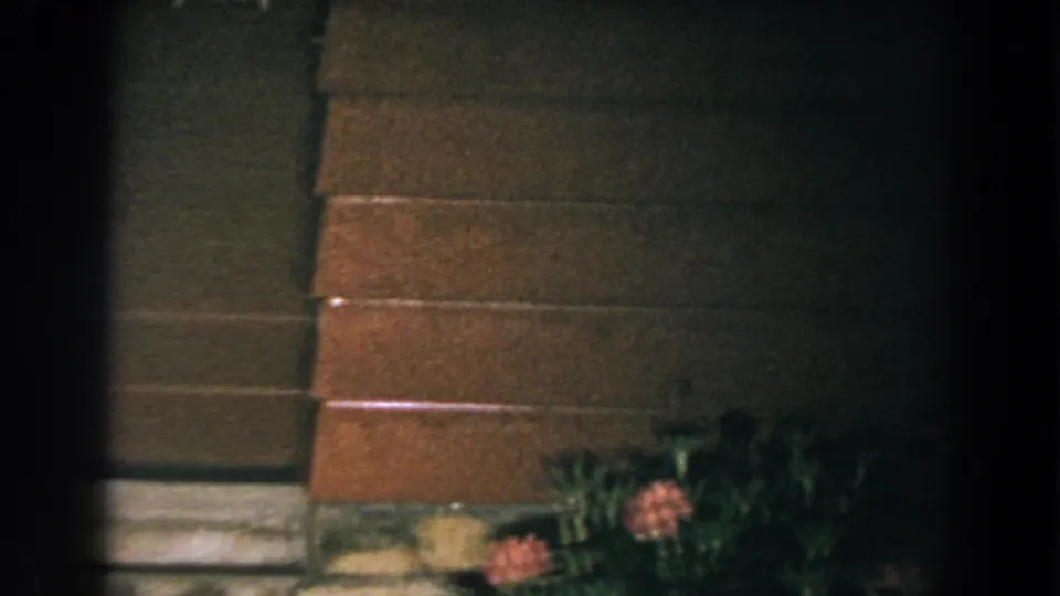 1960 Brighton, Michigan home-movie frame with exterior steps, siding, and flowers near the house at night.